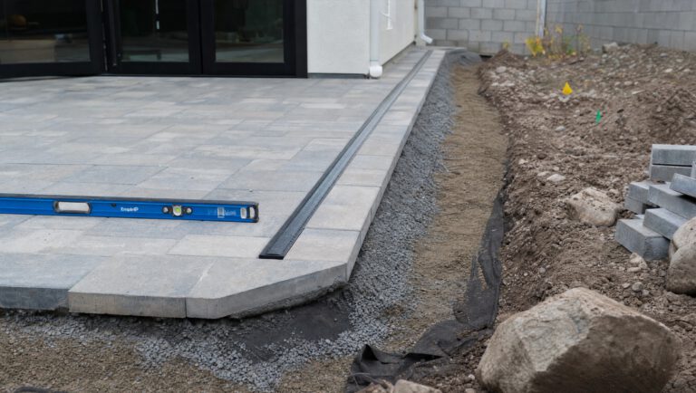 Installation detail of a paver patio edge showing a channel drain and a layered gravel base trench
